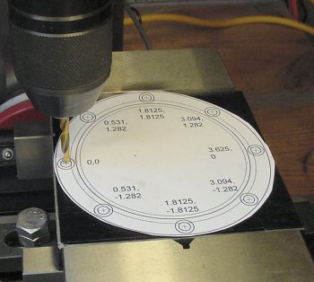 Bolt holes are drilled on a plate in a vise while referencing a paper template.